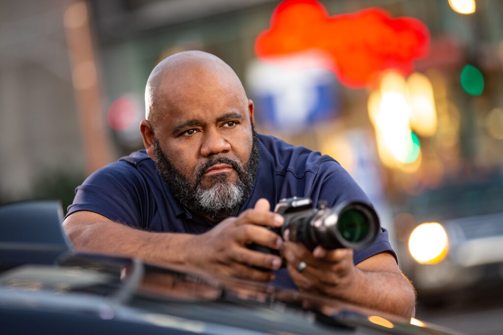 A brown man with a large bald head and thick salt and pepper beard pouts wryly as he rests his camera against his car roof. Red and amber lights blur into the background.
