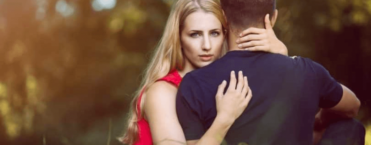 Against a picturesque nature background of golden and green, a gorgeous white blonde woman looks smolderingly at you whilst cradling a dark haired white man, of whom we only see the back.